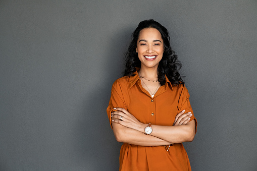 Portrait of a young latin woman with pleasant smile and crossed arms isolated on grey wall with copy space. Cheerful hispanic woman on grey background with copy space. Beautiful girl with folded arms looking at camera against grey wall.